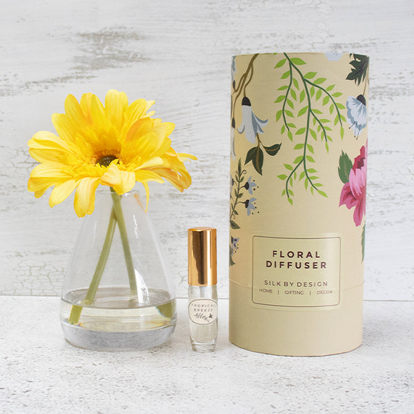 A floral diffuser set featuring a small glass bottle with a metallic gold cap next to its tall beige cylindrical packaging tube decorated with green leaves and colorful flowers. A yellow daisy is placed in a clear glass vase on a light textured surface against a white rustic background.